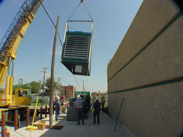 Generator enclosure crane-lifted against concrete block building wall with workers guiding placement