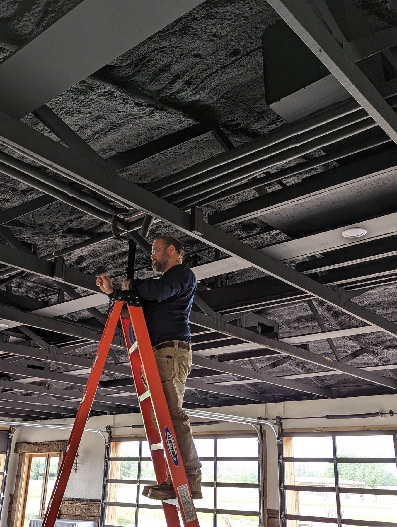 Hartzell's Heat and Air technician on a ladder installing ductwork between the ceiling trusses at The Cedar Gate event venue near Loyal, Oklahoma