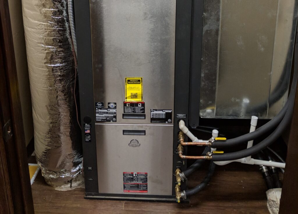 ClimateMaster commercial geothermal water-source heat pump cabinet with copper loop supply and return lines, brass isolation valves, and insulated ground-loop hoses in a mechanical room at the Wheeler Brothers facility in Watonga, Oklahoma, serviced by Hartzell's Heat and Air