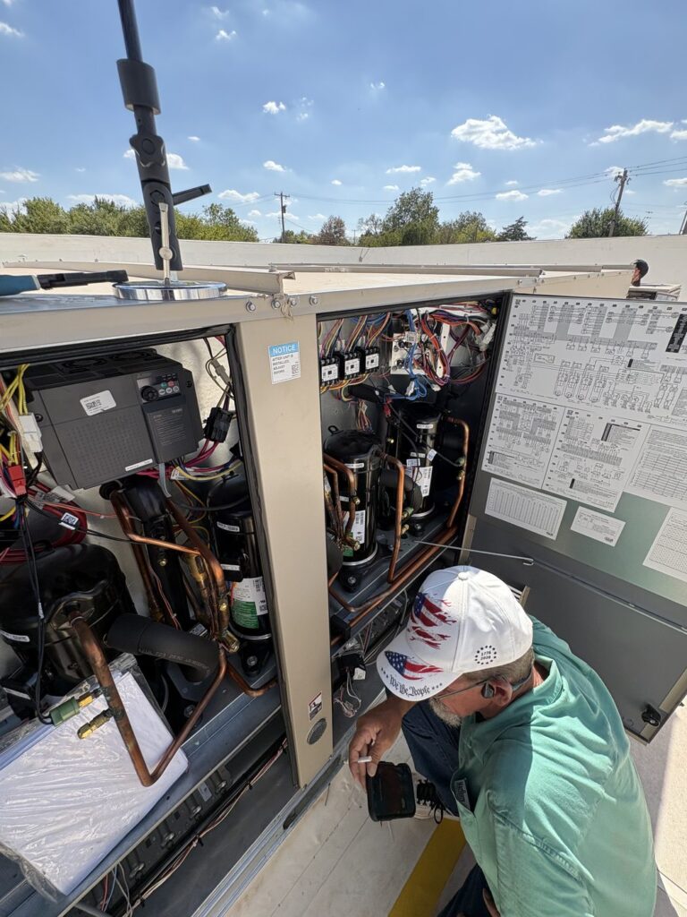 Technician in a white hard hat working inside an open commercial rooftop package unit under a blue sky