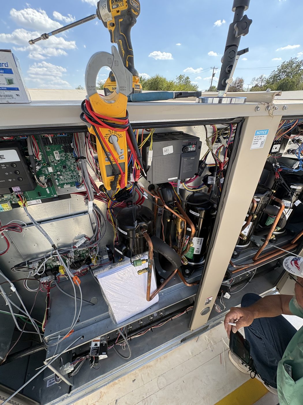 A clamp meter hanging in the opened control section of a commercial rooftop unit, with circuit boards, compressor and copper refrigerant lines visible