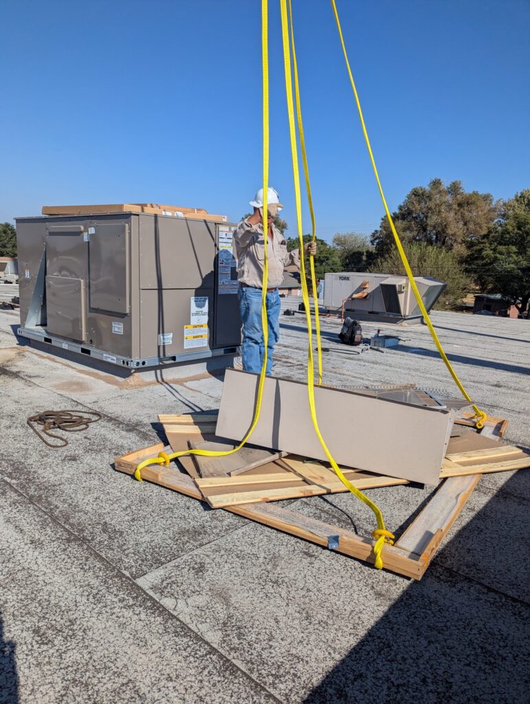 New commercial rooftop HVAC unit being lifted by crane with yellow rigging straps toward a flat roof